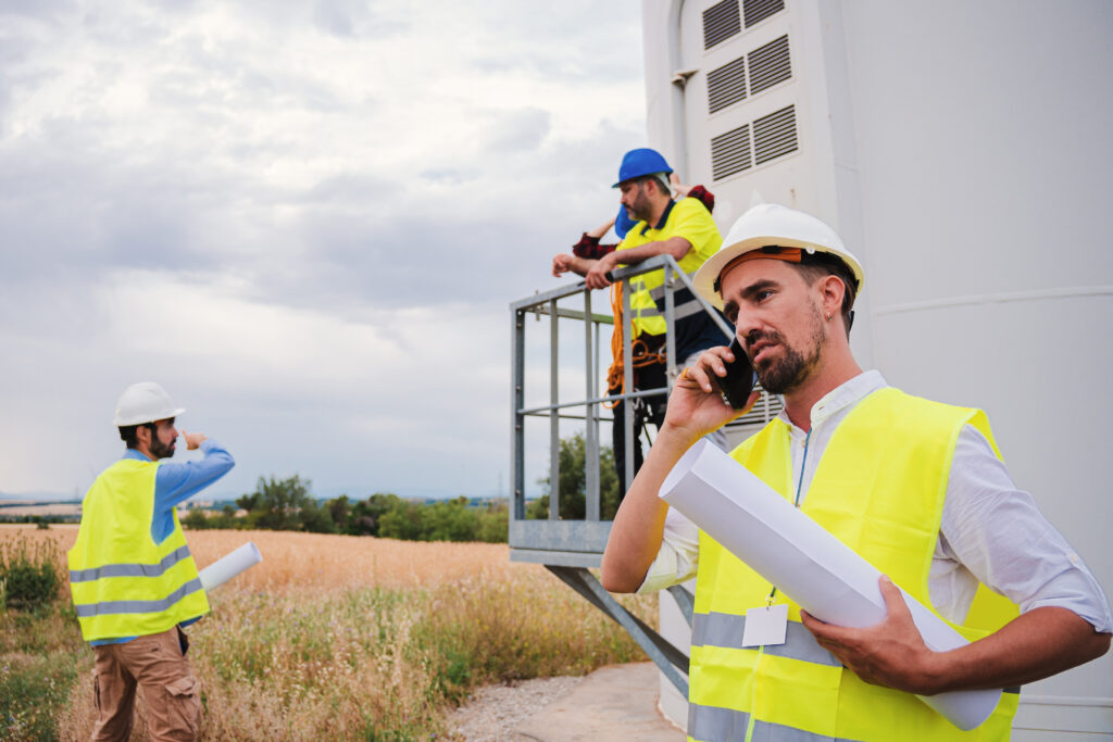 On foreground a caucasian engineer talkin by cell phone holding a blueprint, at background a group of technicians working. One industrial contractor having a smartphone call speaking about the project. High quality photo