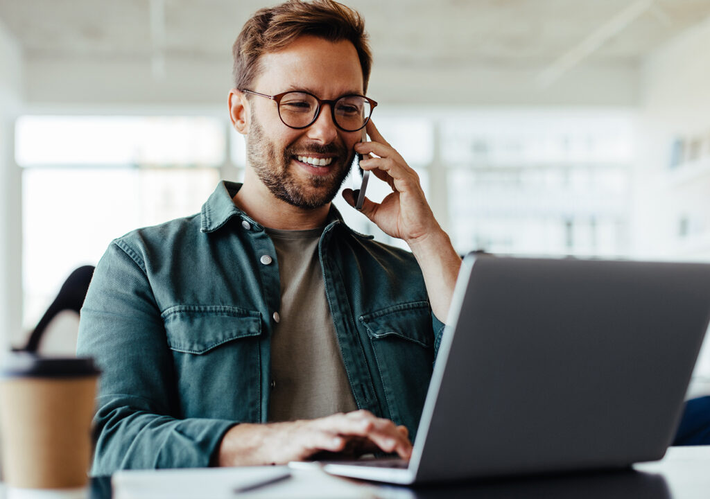 Software designer discussing a new project with his client on the phone. Creative business man working on a laptop in an open plan office.